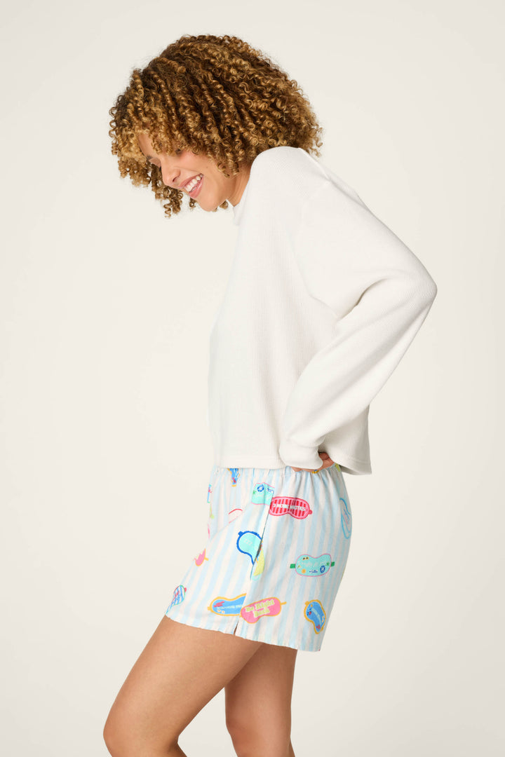A smiling person with curly hair stands in profile against a plain light background, wearing the P.J. Salvage White Long Sleeve Top and colorful shorts featuring a flip-flop print.