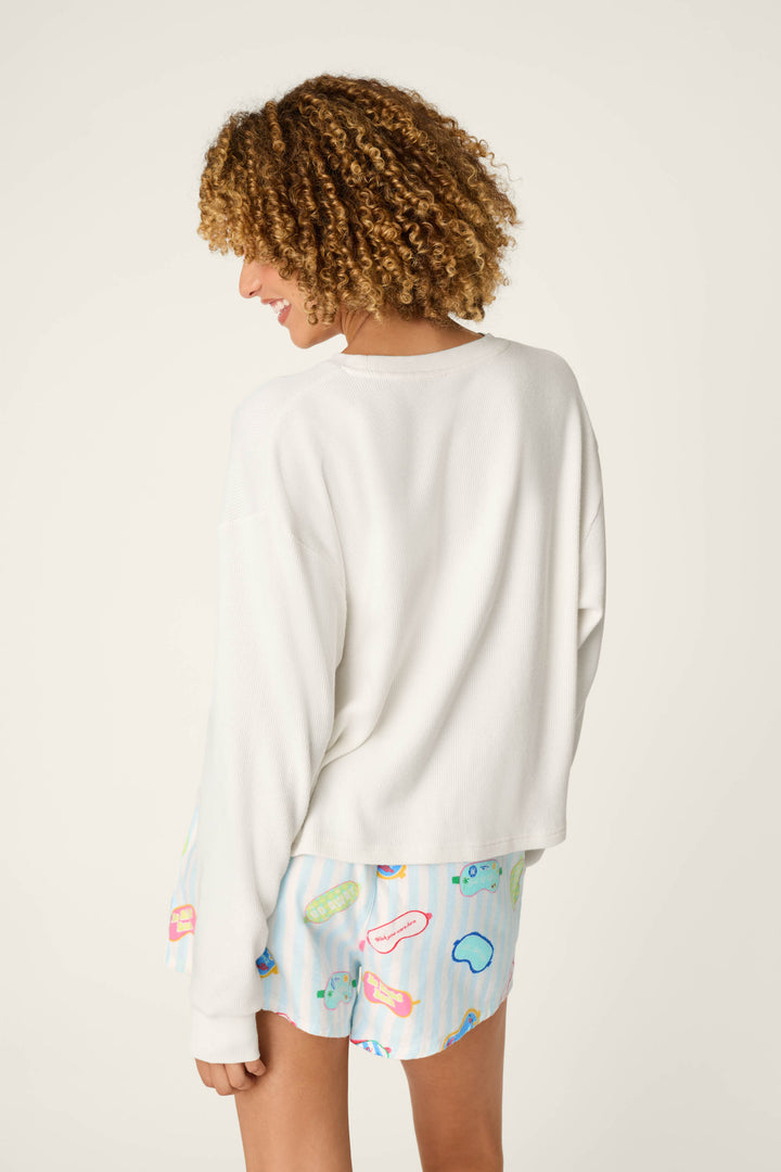 A person with curly hair wears the P.J. Salvage White Long Sleeve Top and colorful printed shorts, standing with their back to the camera against a plain light background.