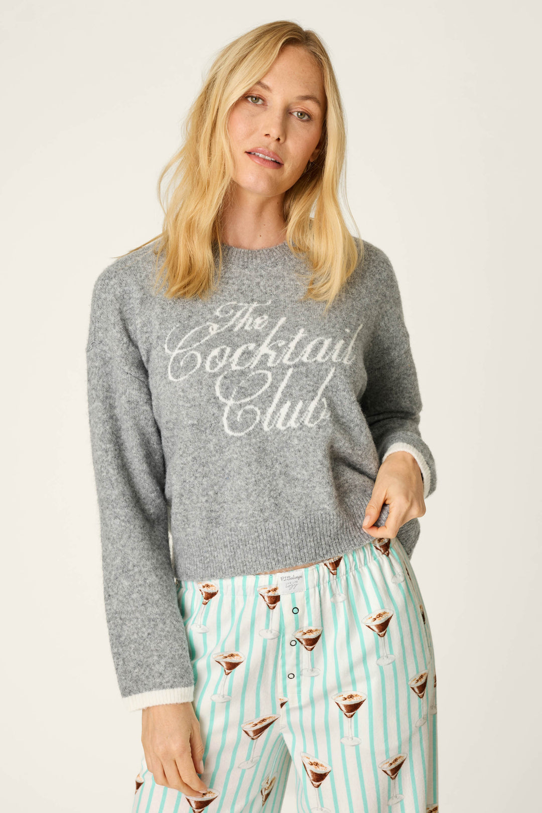 A woman with blonde hair wears the P.J. Salvage The Cocktail Club Snuggle Sweater and striped pajama pants featuring a martini glass print, standing against a plain background.