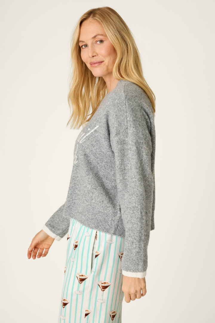 A woman with long blonde hair wears the P.J. Salvage “The Cocktail Club Snuggle Sweater” in gray, paired with white pajama pants featuring a cup-and-stripe pattern. She stands sideways and smiles softly at the camera against a plain light background.
