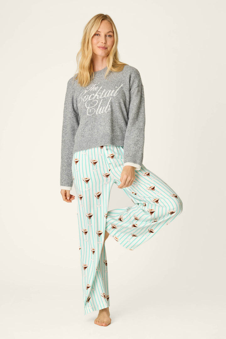 A woman stands barefoot on one leg, wearing the P.J. Salvage The Cocktail Club Snuggle Sweater and light blue pajama pants with a martini glass pattern on white and blue stripes.