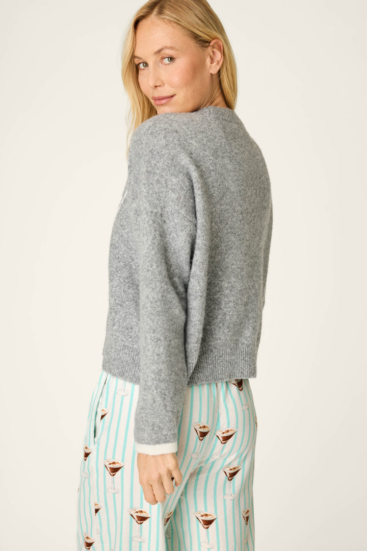 Wearing the P.J. Salvage The Cocktail Club Snuggle Sweater, a person with blonde hair pairs it with white pajama pants featuring teal stripes and coffee cup prints, glancing over their shoulder with a subtle smile.