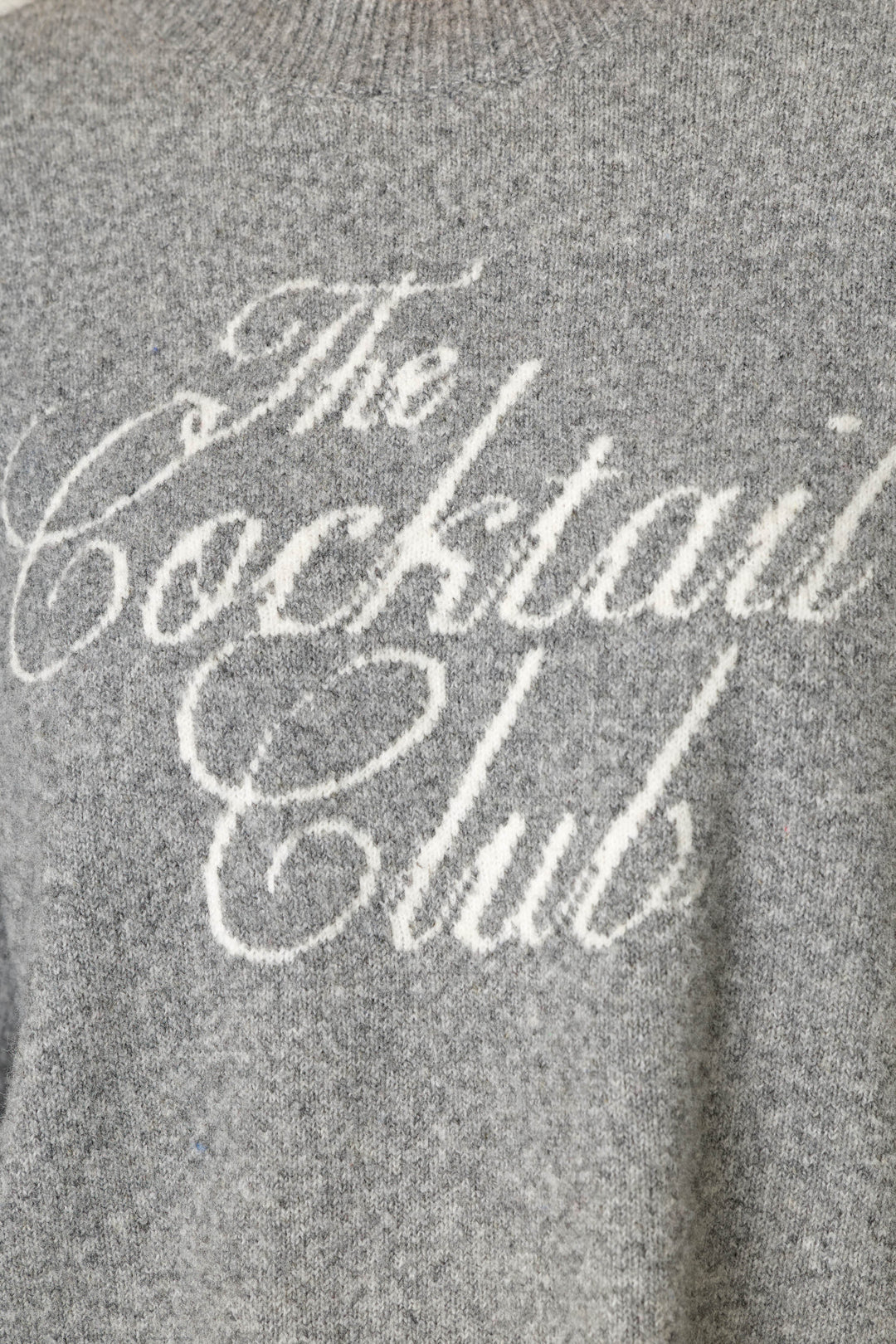 A close-up of the P.J. Salvage The Cocktail Club Snuggle Sweater, a gray knitted sweater featuring “The Cocktail Club” written in white cursive text on the front.