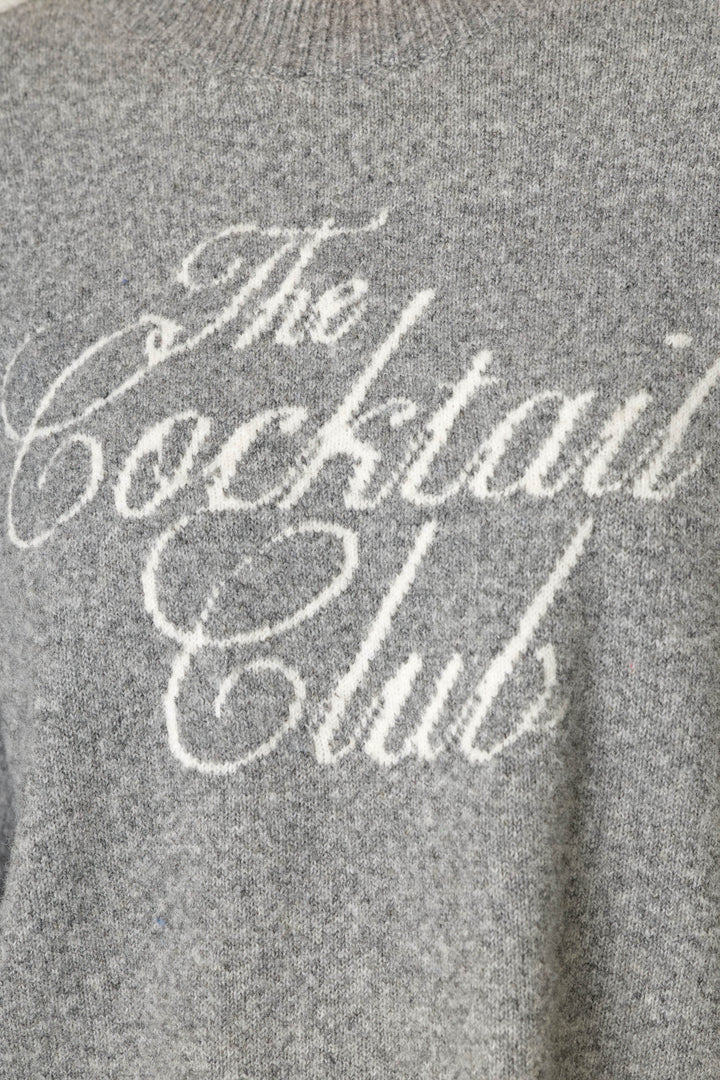 A close-up of the P.J. Salvage The Cocktail Club Snuggle Sweater, a gray knitted sweater featuring “The Cocktail Club” written in white cursive text on the front.