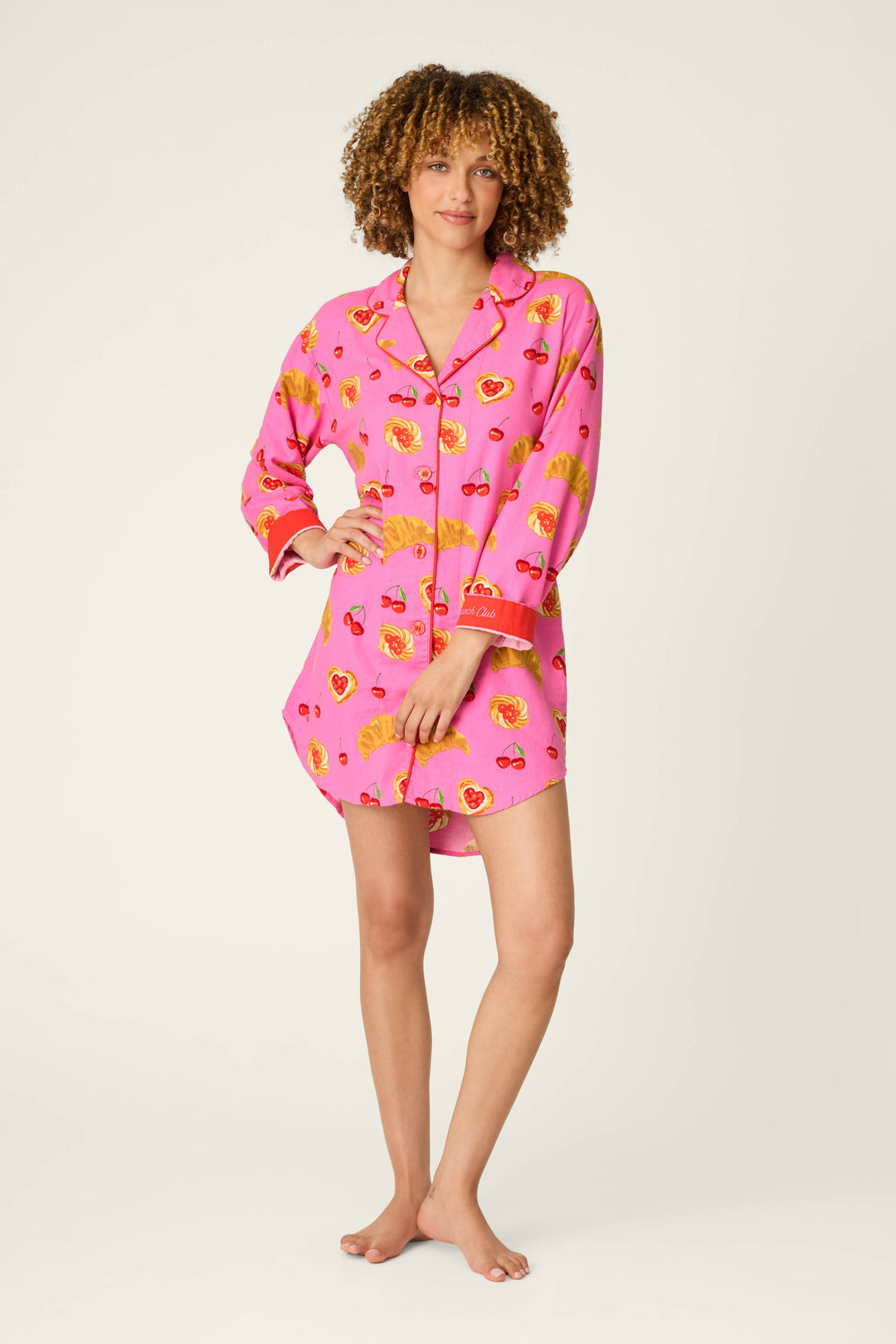 A person with curly hair and a relaxed posture stands barefoot against a plain light background, wearing the P.J. Salvage Sweet Cherry Tart Print Nightshirt—a pink, long-sleeve pajama shirt adorned with playful cherry tart motifs.
