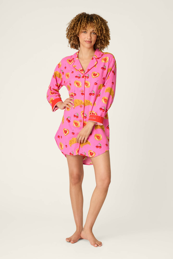 A person with curly hair and a relaxed posture stands barefoot against a plain light background, wearing the P.J. Salvage Sweet Cherry Tart Print Nightshirt—a pink, long-sleeve pajama shirt adorned with playful cherry tart motifs.
