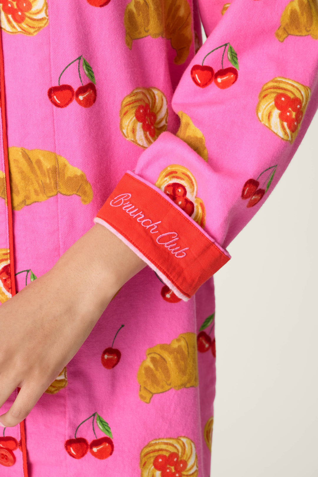 Clad in P.J. Salvage’s Sweet Cherry Tart Print Nightshirt adorned with playful croissant and cherry graphics, a person reveals just their arm and hand. The bright pink shirt features a red cuff embroidered in white script with the words “Brunch Club.”.