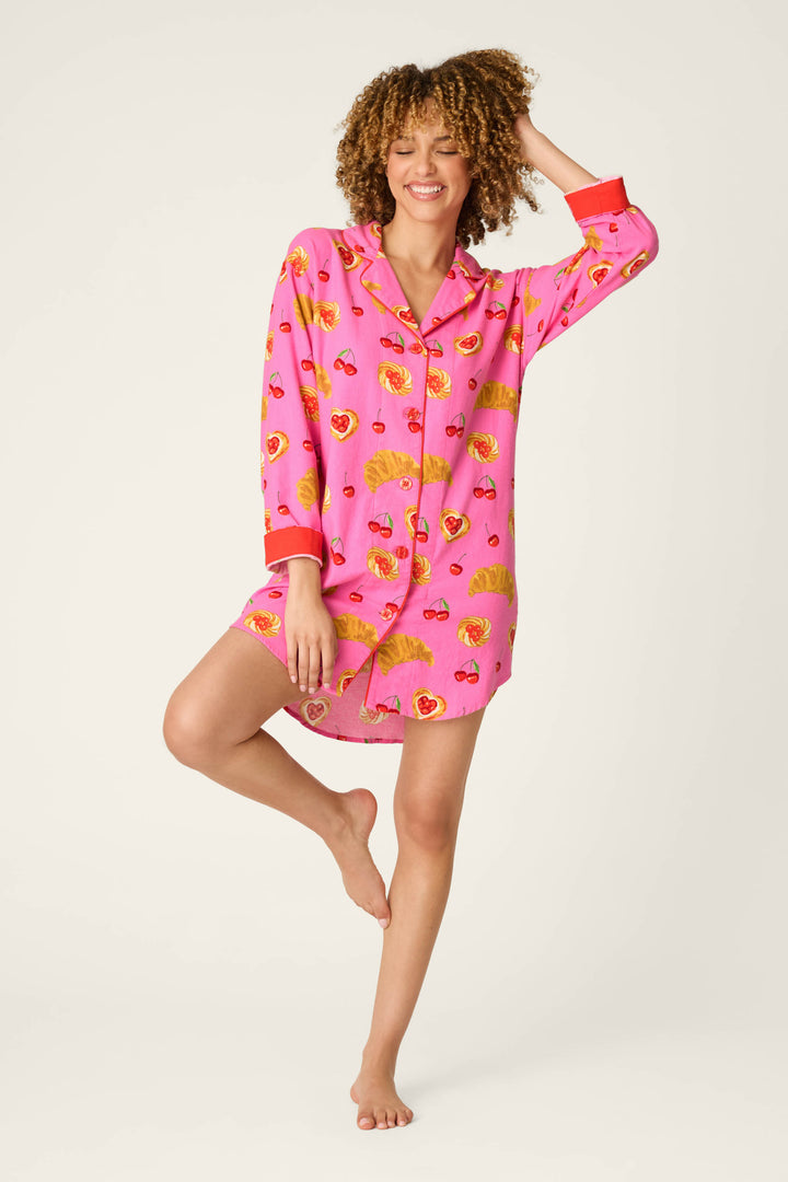 A smiling person with curly hair wears the P.J. Salvage Sweet Cherry Tart Print Nightshirt, standing barefoot on one leg with one arm raised against a plain light background.