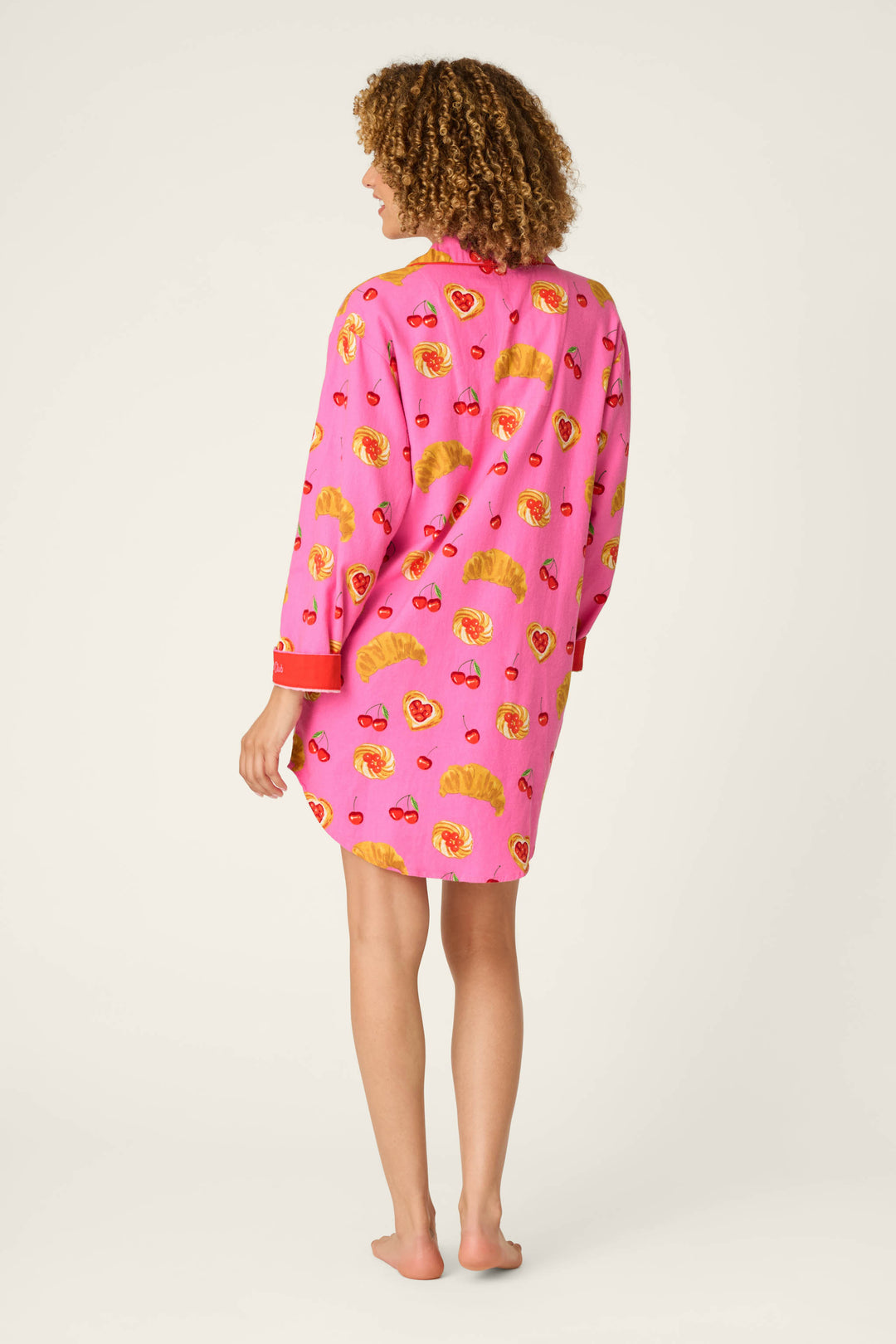 A person with curly hair stands barefoot, facing away, wearing the P.J. Salvage Sweet Cherry Tart Print Nightshirt—a pink long-sleeve shirt featuring a playful pattern of yellow fans, hearts, and cherries—against a plain light background.