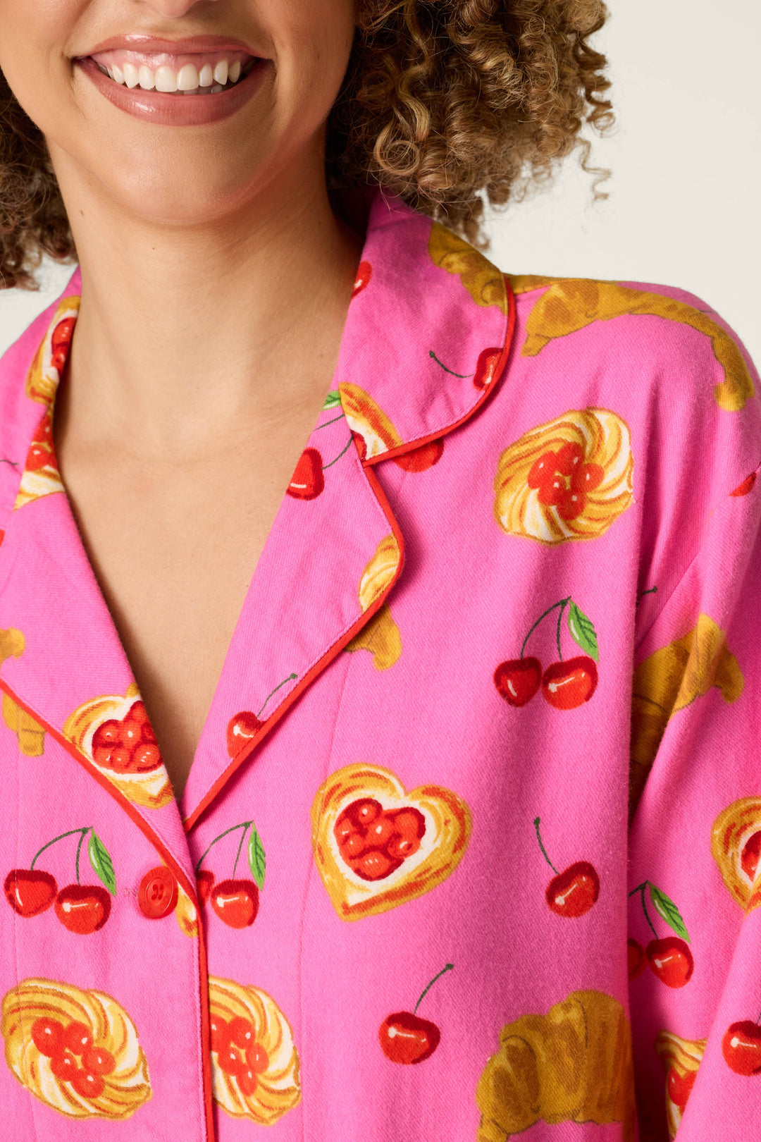 A woman smiles, wearing the P.J. Salvage Sweet Cherry Tart Print Nightshirt. The bright pink nightshirt features a playful pattern of cherries, heart-shaped pastries, and croissants. Only her lower face, neck, and upper torso are visible.