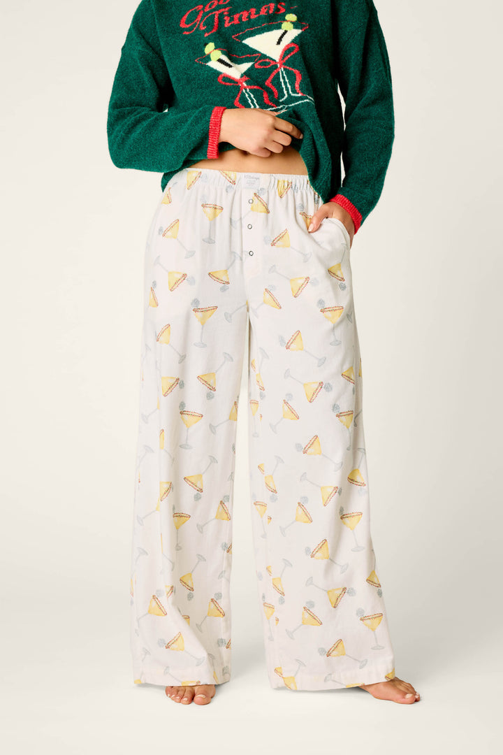 A person wearing P.J. Salvage Happy Hour Cocktail Print Flannel Pajama Pants featuring a margarita glass design and a green Christmas sweater with reindeer antlers, standing barefoot against a plain background.