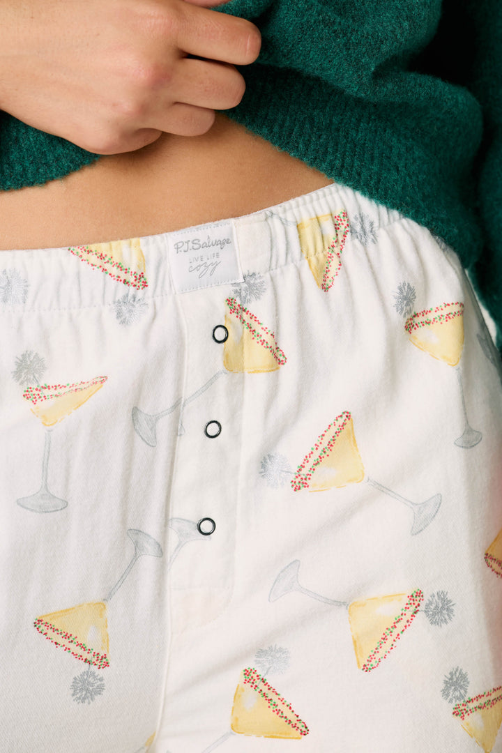 A close-up of a person wearing the P.J. Salvage Happy Hour Cocktail Print Flannel Pajama Pants, featuring a festive cocktail glass pattern and shown with a green sweater, highlighting the waistband and part of their hand.