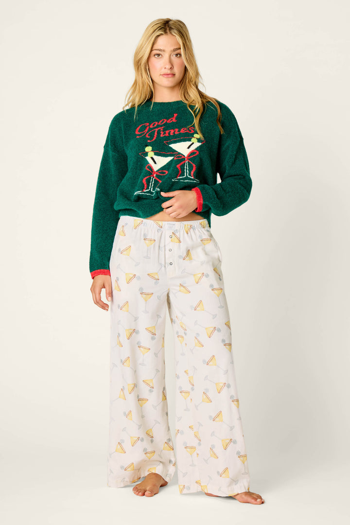 A woman with long blonde hair and a neutral expression stands barefoot wearing P.J. Salvage Happy Hour Cocktail Print Flannel Pajama Pants and a green sweater that says “Good Times” with martini glasses.