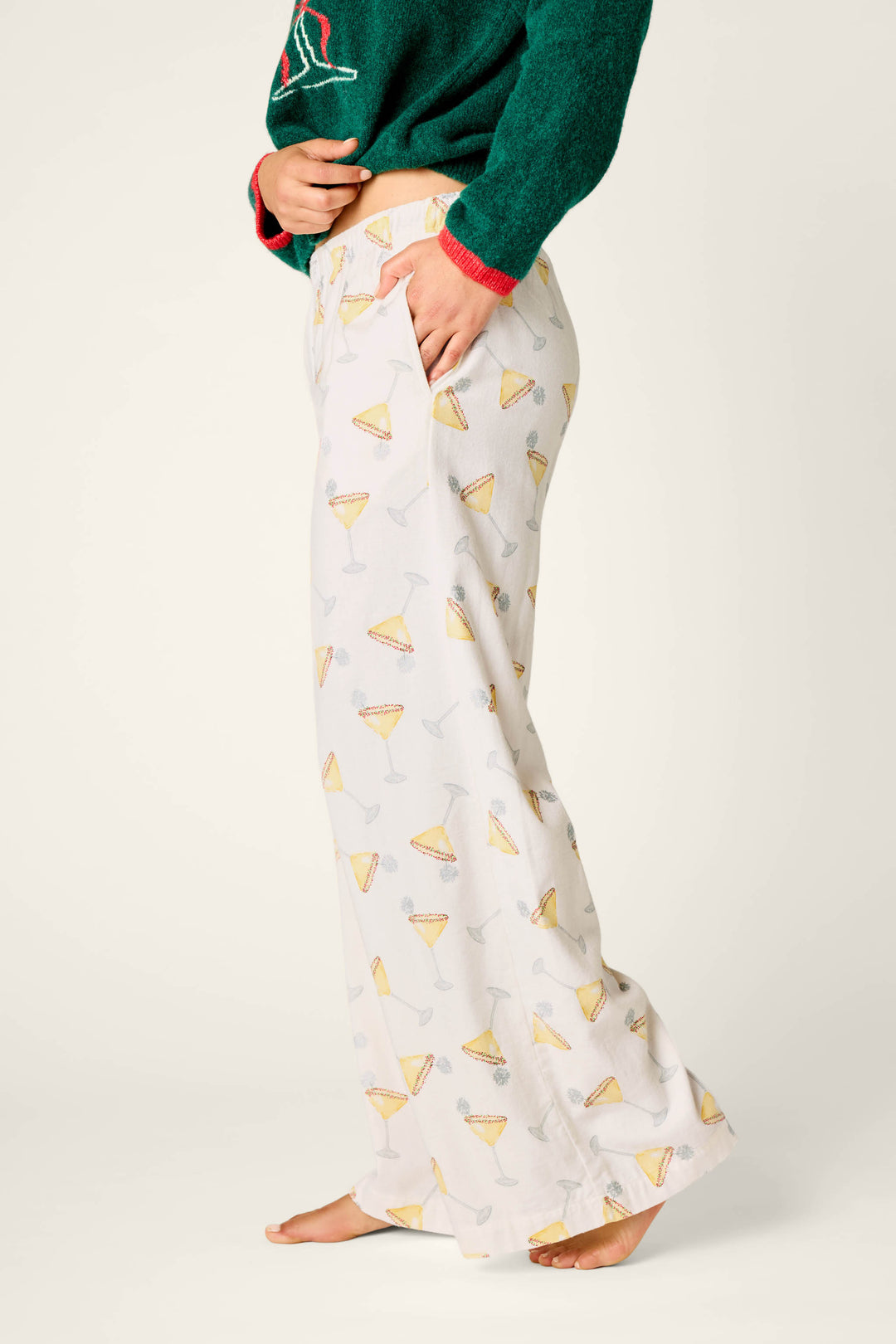 A person wearing P.J. Salvage Happy Hour Cocktail Print Flannel Pajama Pants, featuring a cocktail glass and lemon motif, paired with a green sweater. The individual is barefoot and standing sideways against a plain background.