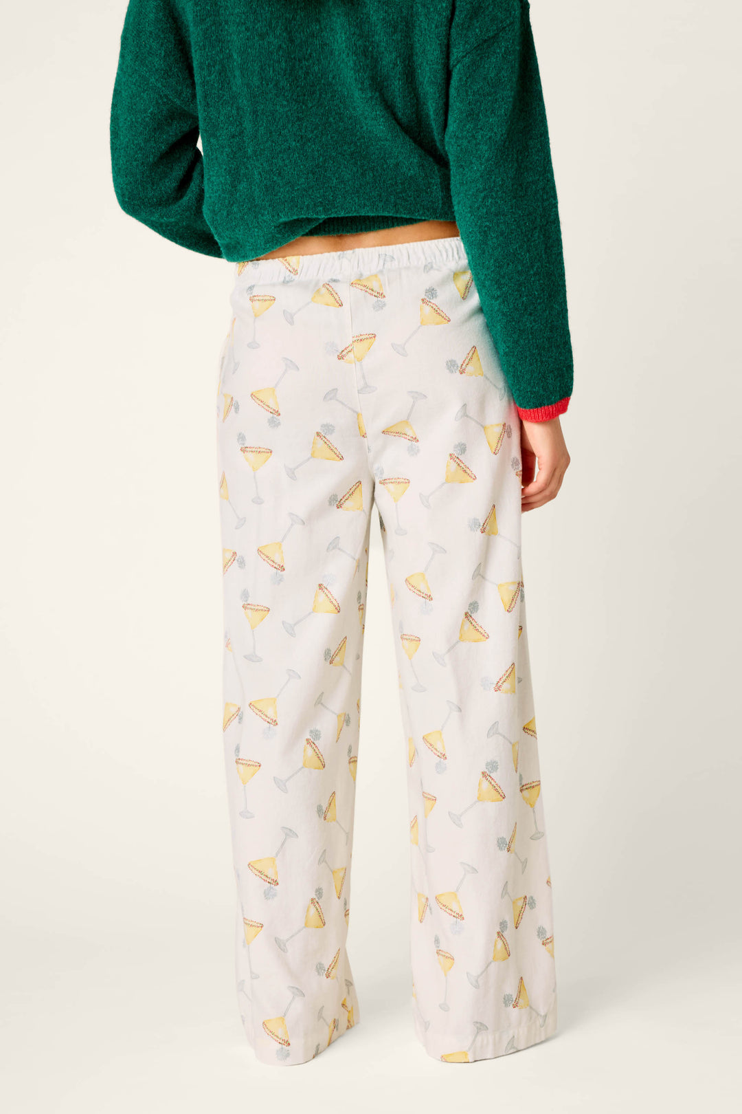 A person wearing a green sweater and P.J. Salvage Happy Hour Cocktail Print Flannel Pajama Pants—featuring a white base with yellow cocktail glass motifs—stands with their back to the camera against a plain background, highlighting the back of the outfit.