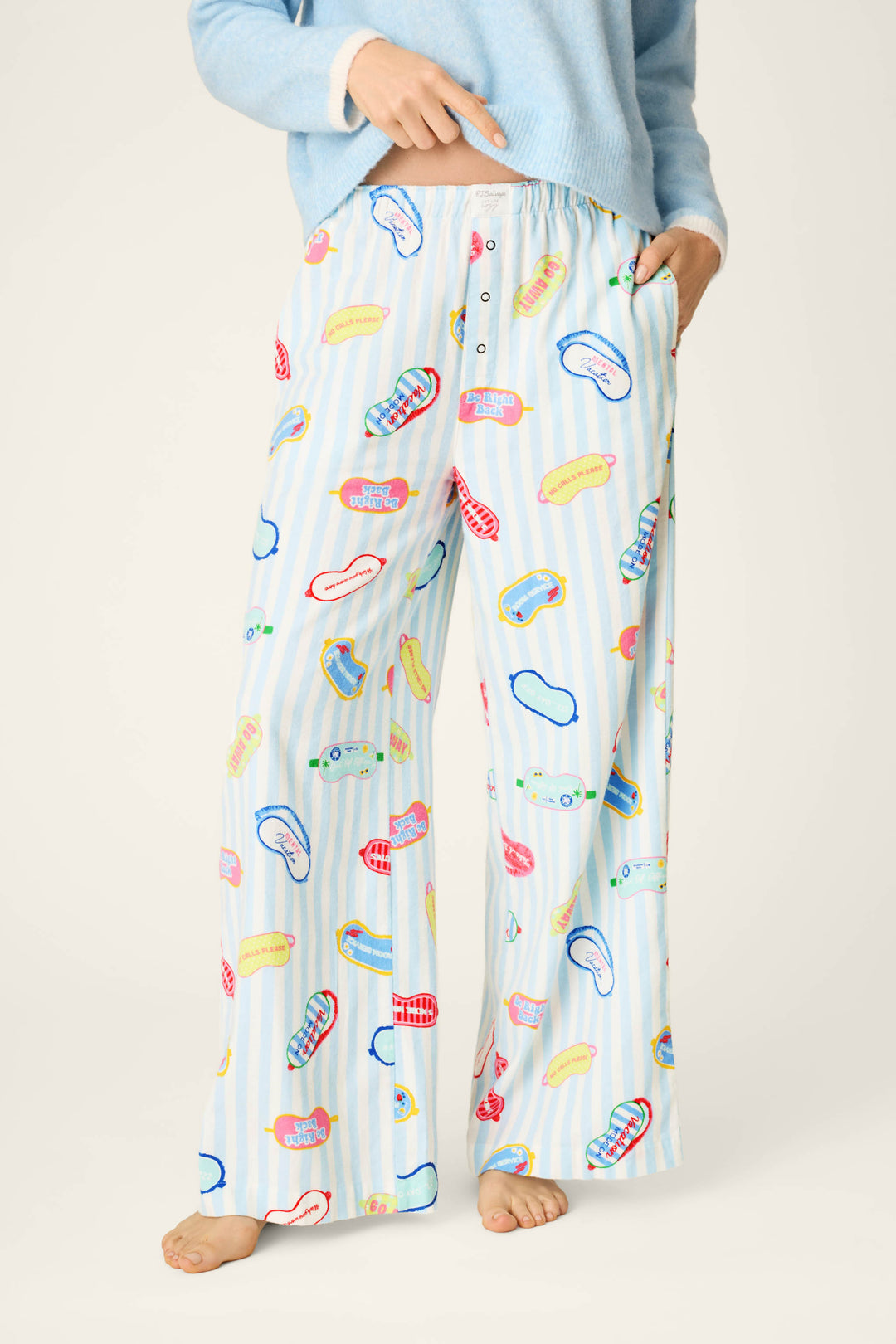 Wearing P.J. Salvage's Out of Office Eyemask Print Flannel Pajama Pants in light blue, this individual is styled with a matching sweater. One hand rests in a pocket while the other points to the pajama pants, with the image capturing their body from the shoulders down.