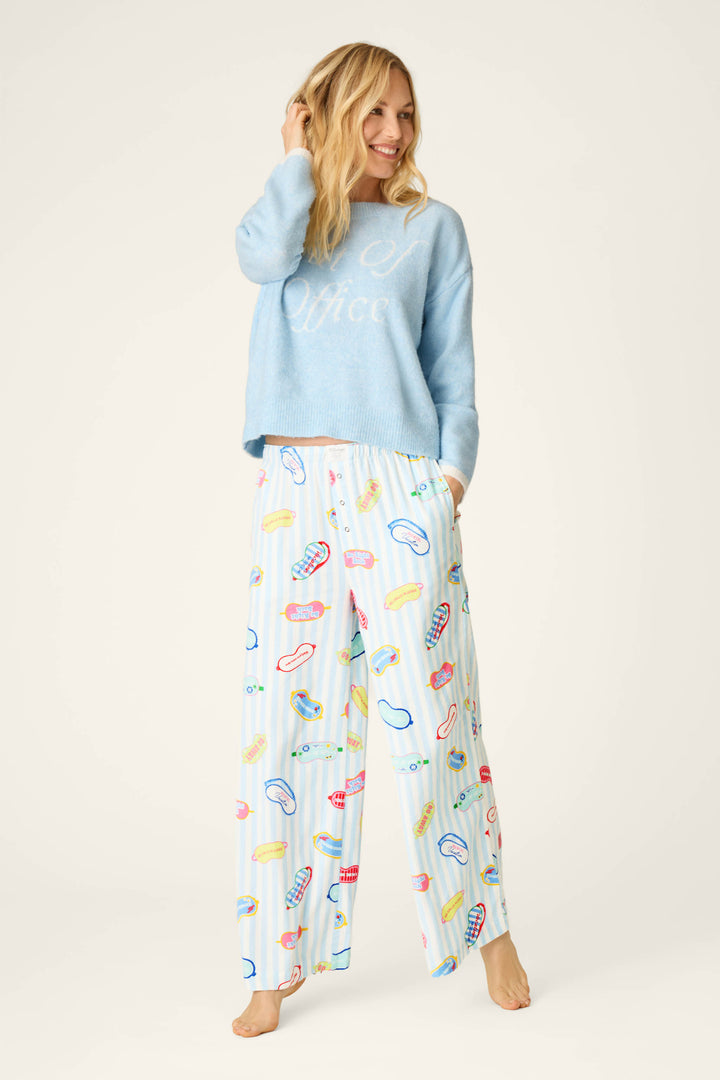 A blonde woman smiles as she wears a light blue sweater with "Out of Office" written on it and the P.J. Salvage Out of Office Eyemask Print Flannel Pajama Pants, which are loose, light blue and white striped pajama pants covered in colorful illustrations of sleep masks and vacation-themed text. She is barefoot.