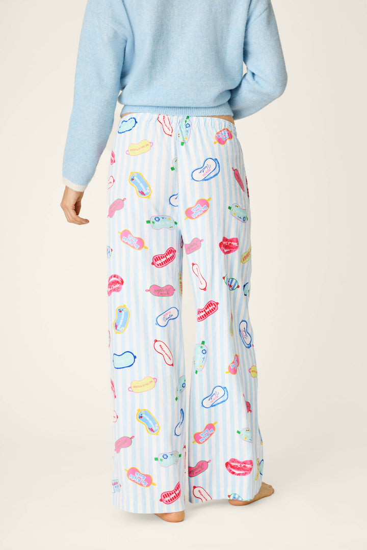 A person stands barefoot and facing away from the camera against a neutral background, wearing a blue sweater paired with P.J. Salvage's Out of Office Eyemask Print Flannel Pajama Pants—loose white pajama pants featuring colorful lip, eye, and sandal patterns.