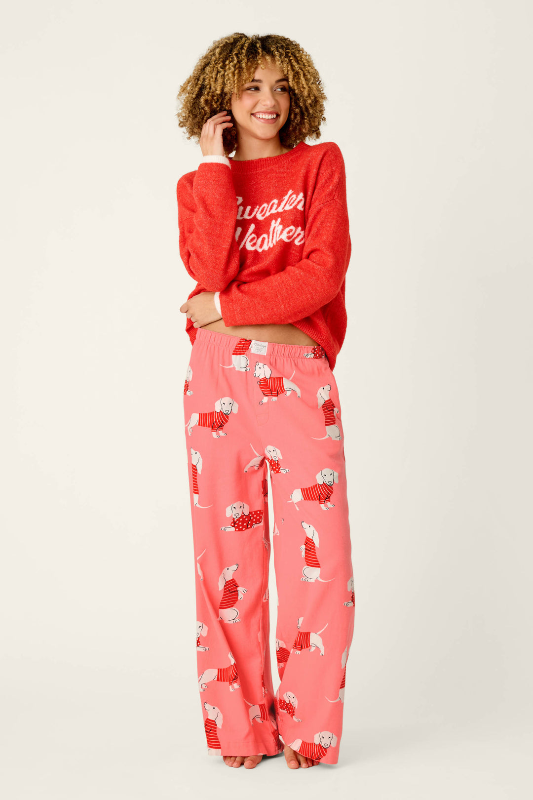 A smiling woman with curly hair wears a red Sweater Weather sweater and P.J. Salvage’s Dachshund Print Flannel Pajama Pants, standing barefoot against a plain light background.