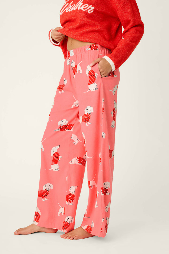 A person stands barefoot against a neutral background, wearing P.J. Salvage Dachshund Print Flannel Pajama Pants featuring playful images of dachshunds in red sweaters; only the lower half of the person is visible.