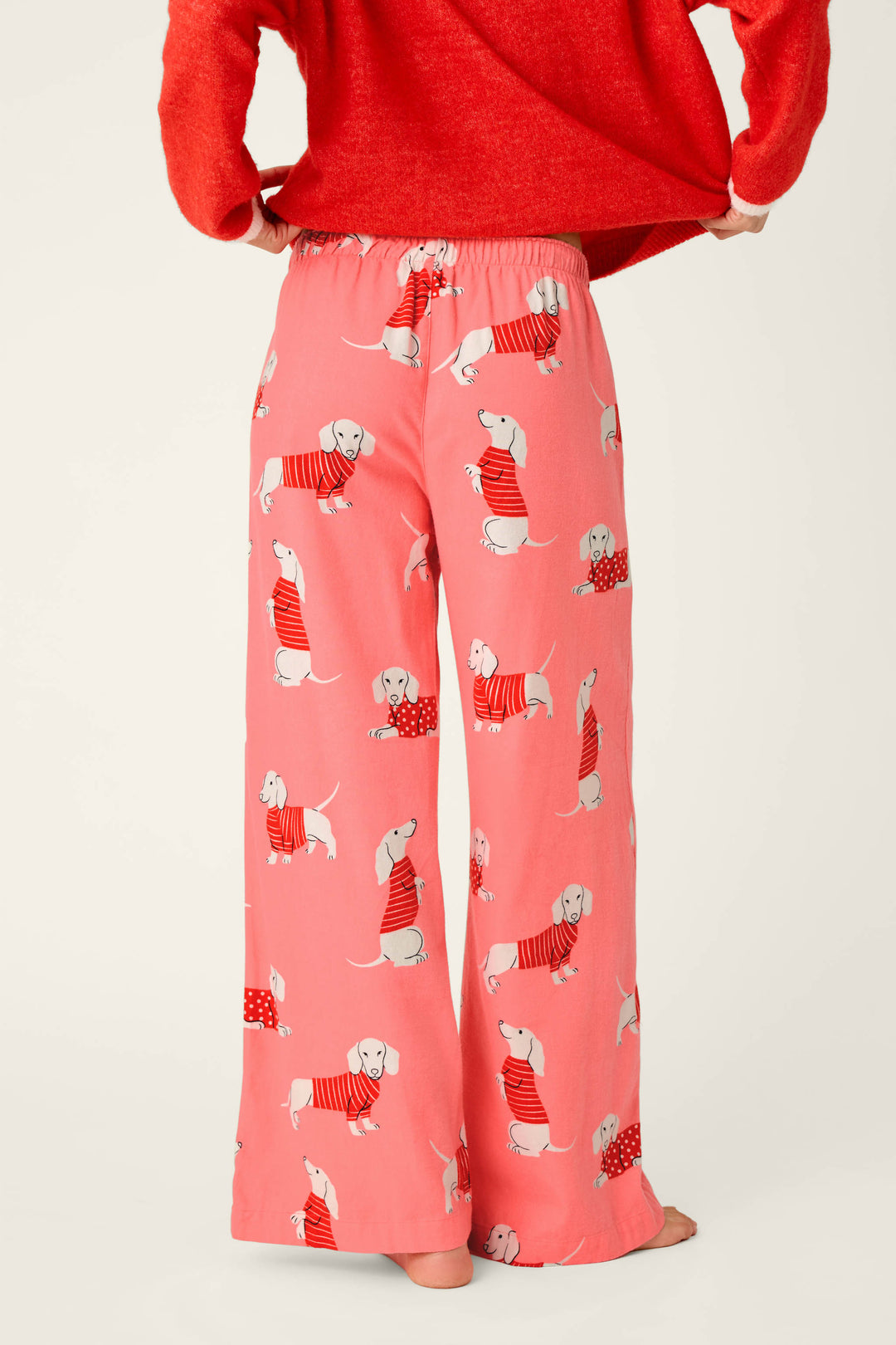 A person is standing barefoot and facing away from the camera, wearing P.J. Salvage Dachshund Print Flannel Pajama Pants in pink paired with a red sweater.