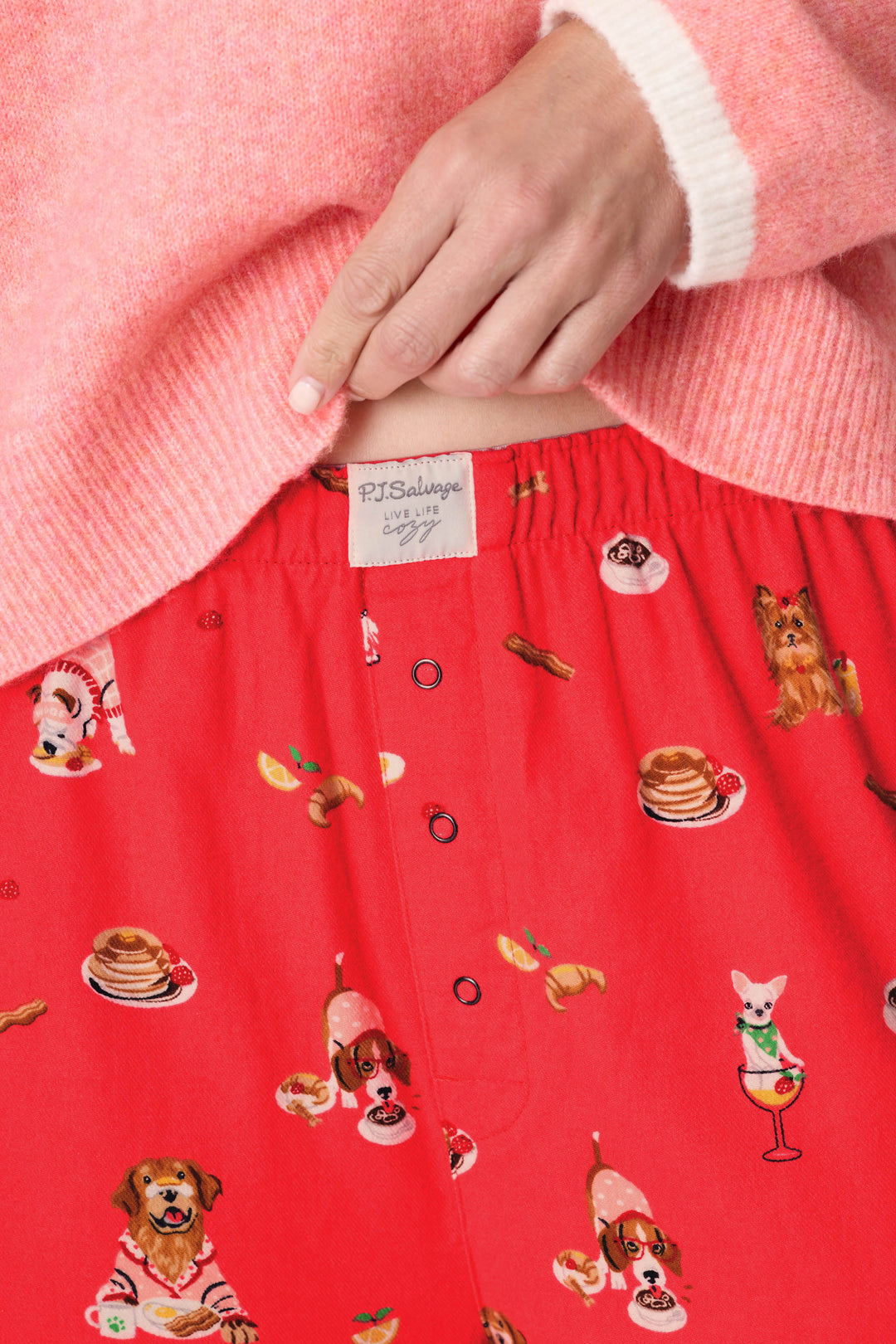 Wearing the P.J. Salvage Dogs Who Brunch Flannel Pajama Pants, which feature a fun dog and breakfast print on a red background, the person pulls at the waistband to reveal the brand’s label. They pair the pajama pants with a pink and white sweater.
