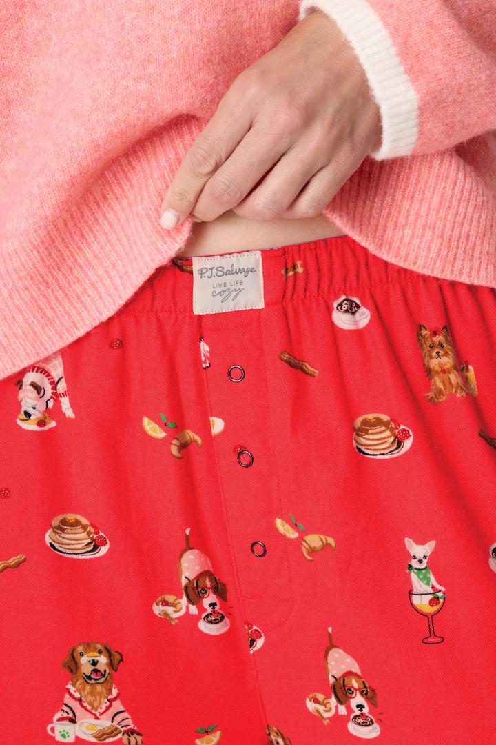 Wearing the P.J. Salvage Dogs Who Brunch Flannel Pajama Pants, which feature a fun dog and breakfast print on a red background, the person pulls at the waistband to reveal the brand’s label. They pair the pajama pants with a pink and white sweater.