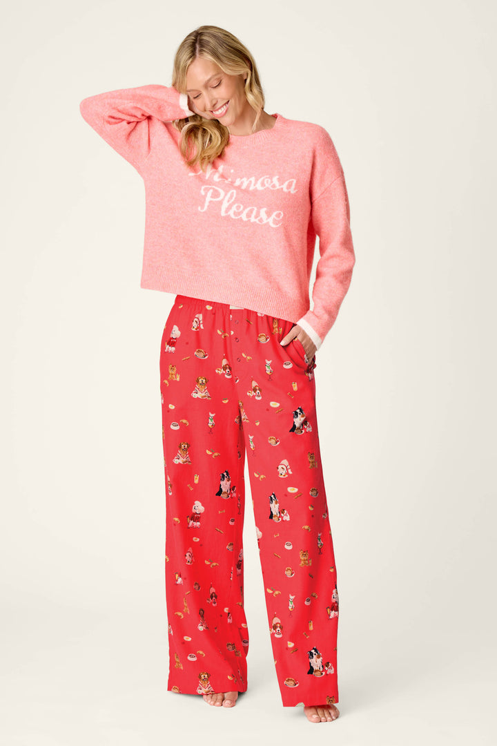 A smiling woman stands against a plain background wearing P.J. Salvage Dogs Who Brunch Flannel Pajama Pants, which are red and decorated with small dog illustrations, paired with a pink sweater featuring “Mimosa Please” text.
