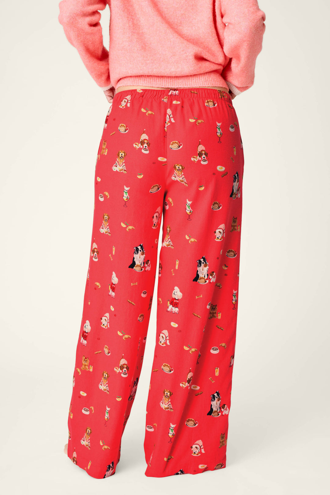 A person is shown from the back wearing P.J. Salvage Dogs Who Brunch Flannel Pajama Pants, featuring a bright red print with cartoon dogs and brunch food. The pants are paired with a coordinating red-pink sweater, against a plain light-colored background.