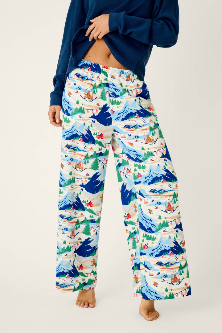 A person is shown standing barefoot against a plain background, wearing P.J. Salvage Ski Ya Later Skiing Bears Print Flannel Pajama Pants featuring a playful mountain, forest, and camping motif, paired with a navy blue sweatshirt. Their face is not visible.