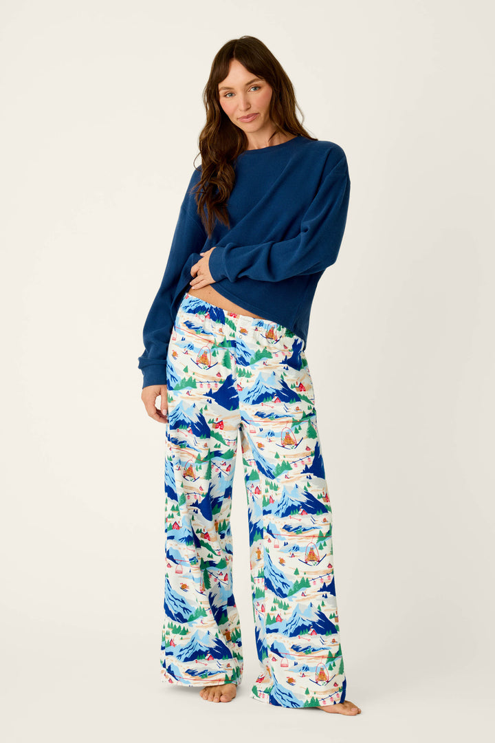 A woman stands barefoot, wearing a blue sweater and P.J. Salvage’s Ski Ya Later Skiing Bears Print Flannel Pajama Pants, which feature a vibrant mountain and landscape design, as she poses against a plain white background.