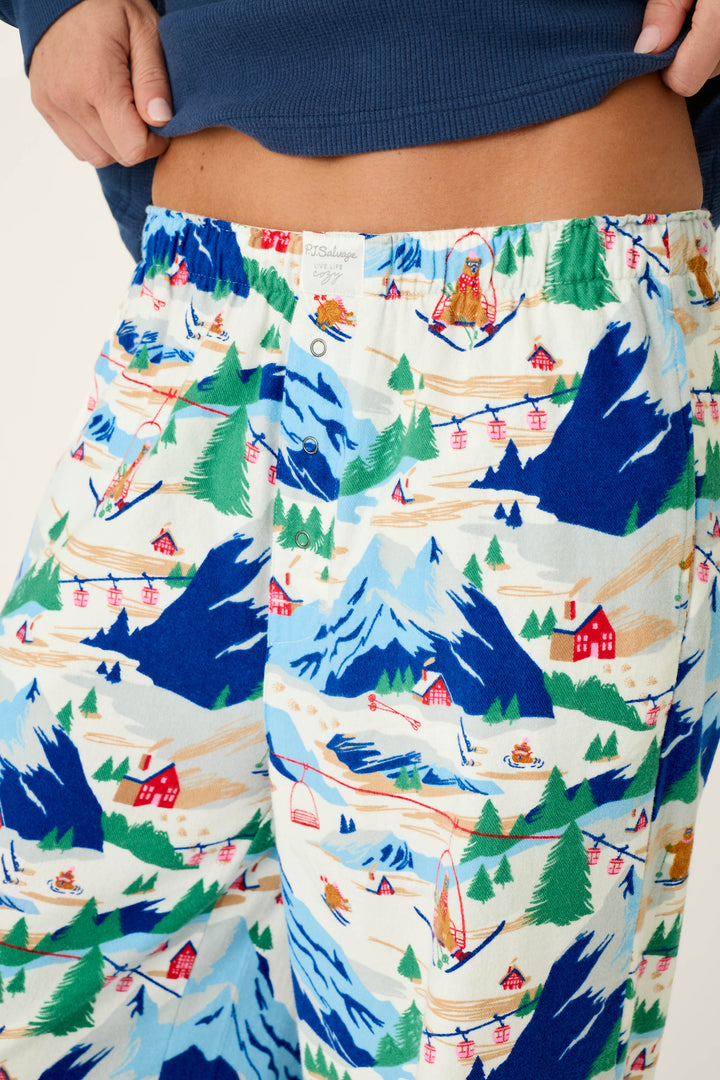 Close-up of a person wearing P.J. Salvage’s Ski Ya Later Skiing Bears Print Flannel Pajama Pants, which feature a colorful mountain ski resort scene with blue mountains, red cable cars, trees, and ski lodges. The person is lifting a navy shirt to reveal the waistband.