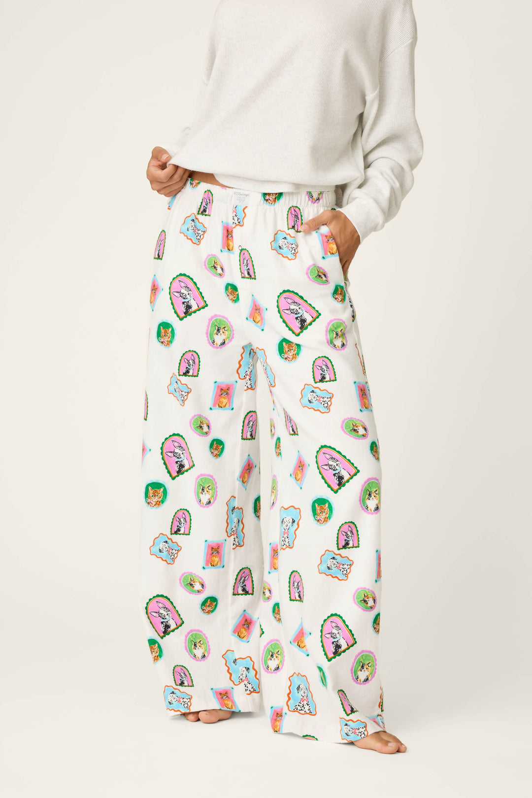 A person wears the P.J. Salvage Picture Purrfect Cat & Dog Portraits Flannel Pajama Pants—loose white pajama pants featuring colorful cartoon-style cat and face prints—paired with a white sweatshirt. The individual’s head is out of frame.