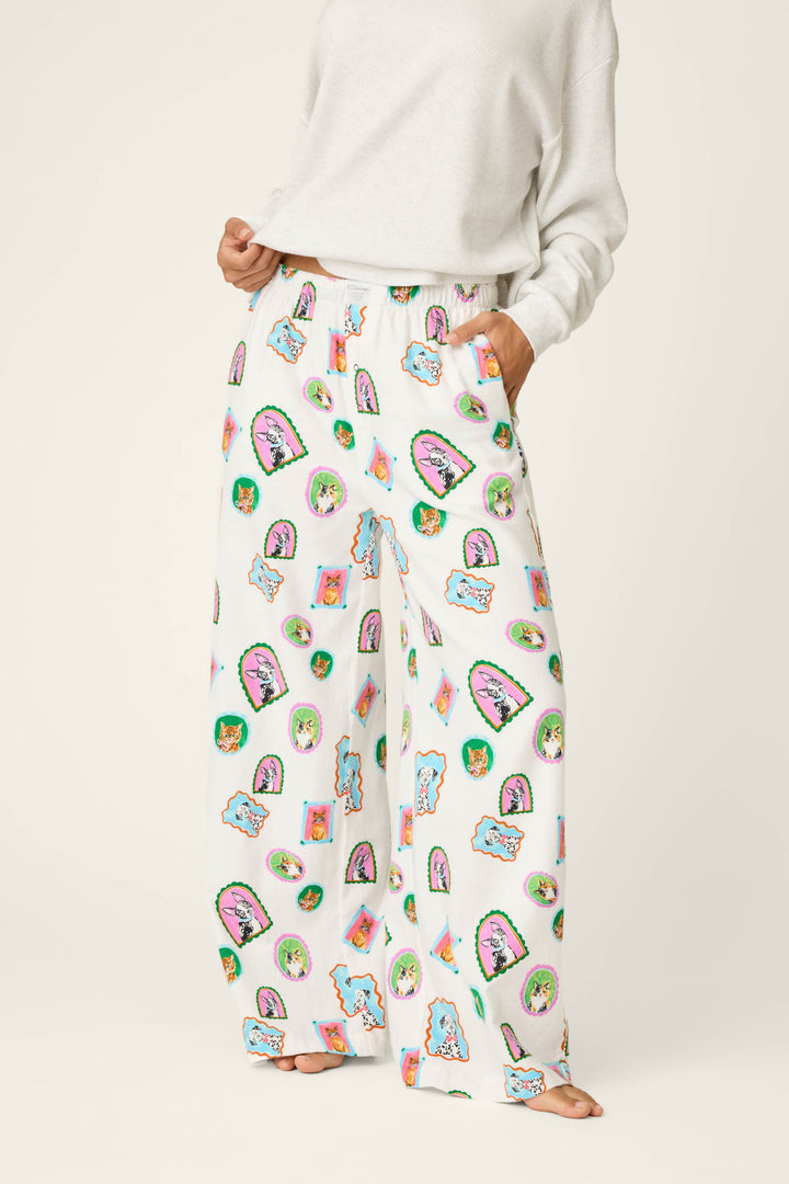 A person wears the P.J. Salvage Picture Purrfect Cat & Dog Portraits Flannel Pajama Pants—loose white pajama pants featuring colorful cartoon-style cat and face prints—paired with a white sweatshirt. The individual’s head is out of frame.