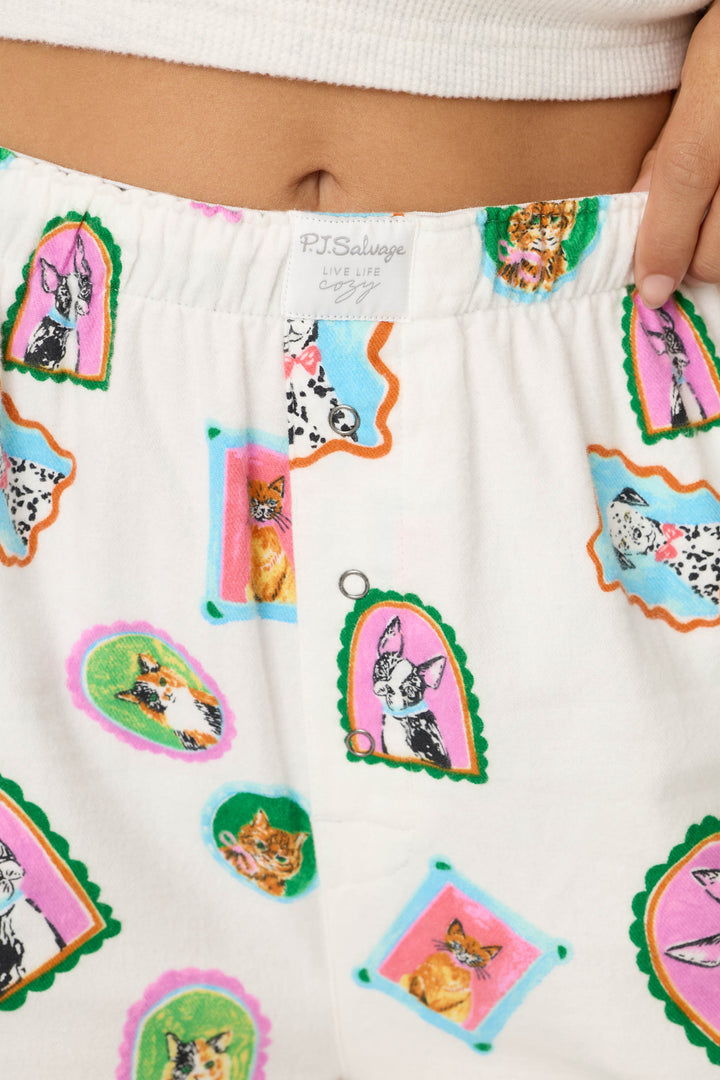 Close-up of a person's waist wearing P.J. Salvage Picture Purrfect Cat & Dog Portraits Flannel Pajama Pants, featuring colorful framed cat and dog illustrations, a visible brand tag, and part of a hand resting on the waistband.
