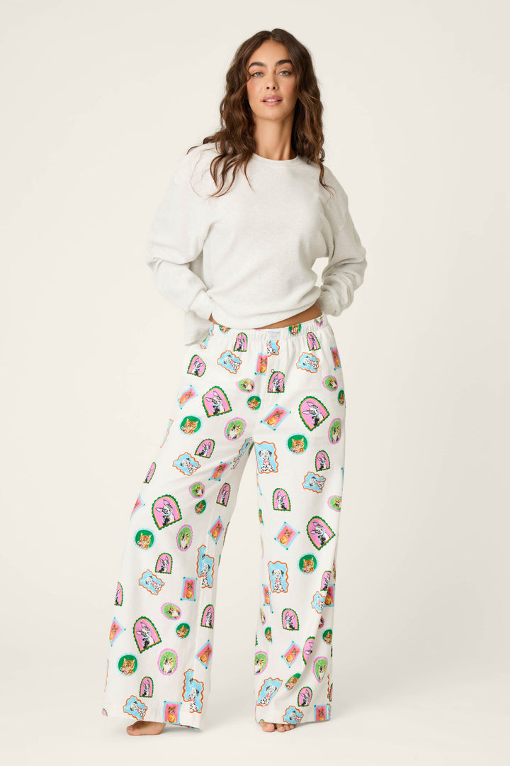 A woman stands barefoot against a plain light background, wearing a white sweater and P.J. Salvage Picture Purrfect Cat & Dog Portraits Flannel Pajama Pants featuring colorful cartoon faces and shapes.