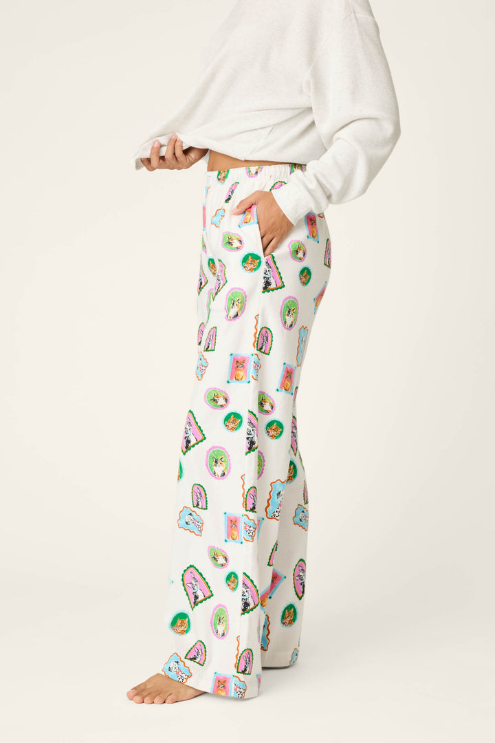 A person stands barefoot against a plain background, hands in pockets, wearing P.J. Salvage's Picture Purrfect Cat & Dog Portraits Flannel Pajama Pants—white lounge pants featuring colorful, playful cat and dog graphics—paired with a white long-sleeve top.