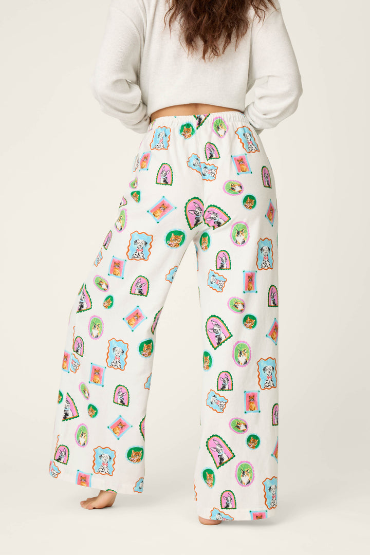 A person stands barefoot, facing away, wearing P.J. Salvage Picture Purrfect Cat & Dog Portraits Flannel Pajama Pants featuring colorful cartoon animal faces, paired with a white cropped long-sleeve top. The background is plain and light-colored.