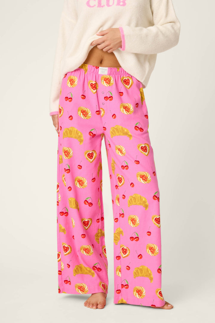A person wearing P.J. Salvage Sweet Cherry Tart Print Pajama Pants, featuring croissant, heart, and cherry motifs, paired with a cream sweater. The person is barefoot and their face is not visible.