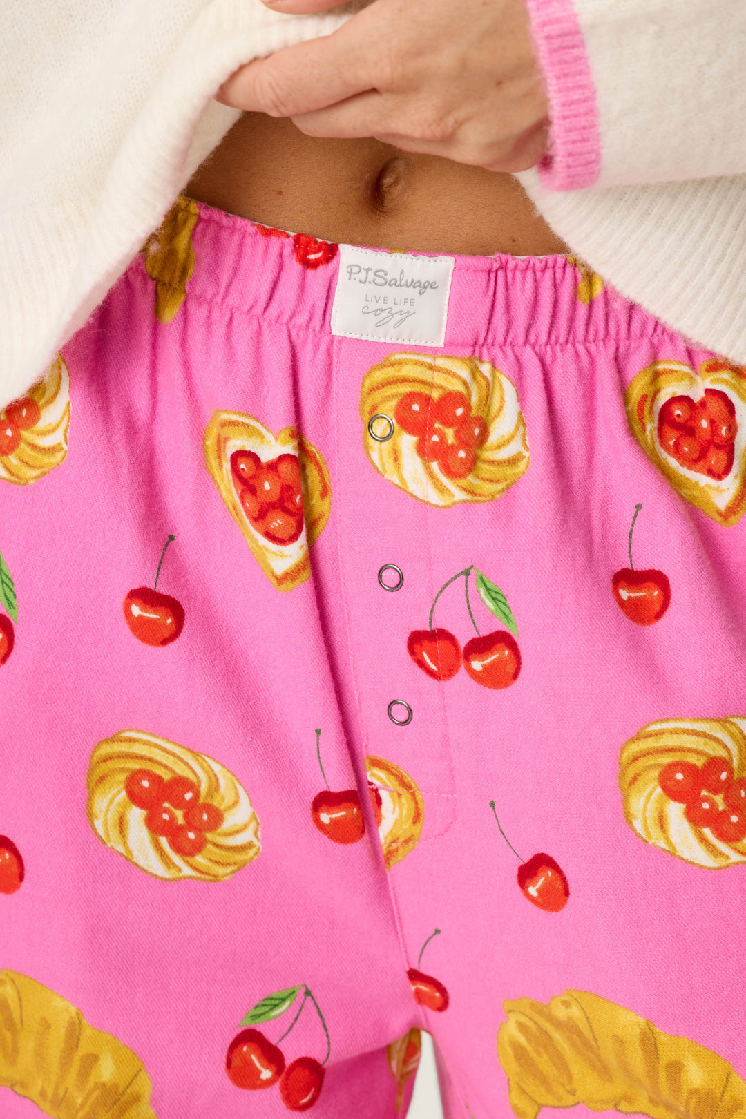 Close-up of a person wearing P.J. Salvage Sweet Cherry Tart Print Pajama Pants, featuring bright pink fabric with cherry and pastry designs, partially covered by a cream sweater. The waistband displays the visible P.J. Salvage label.