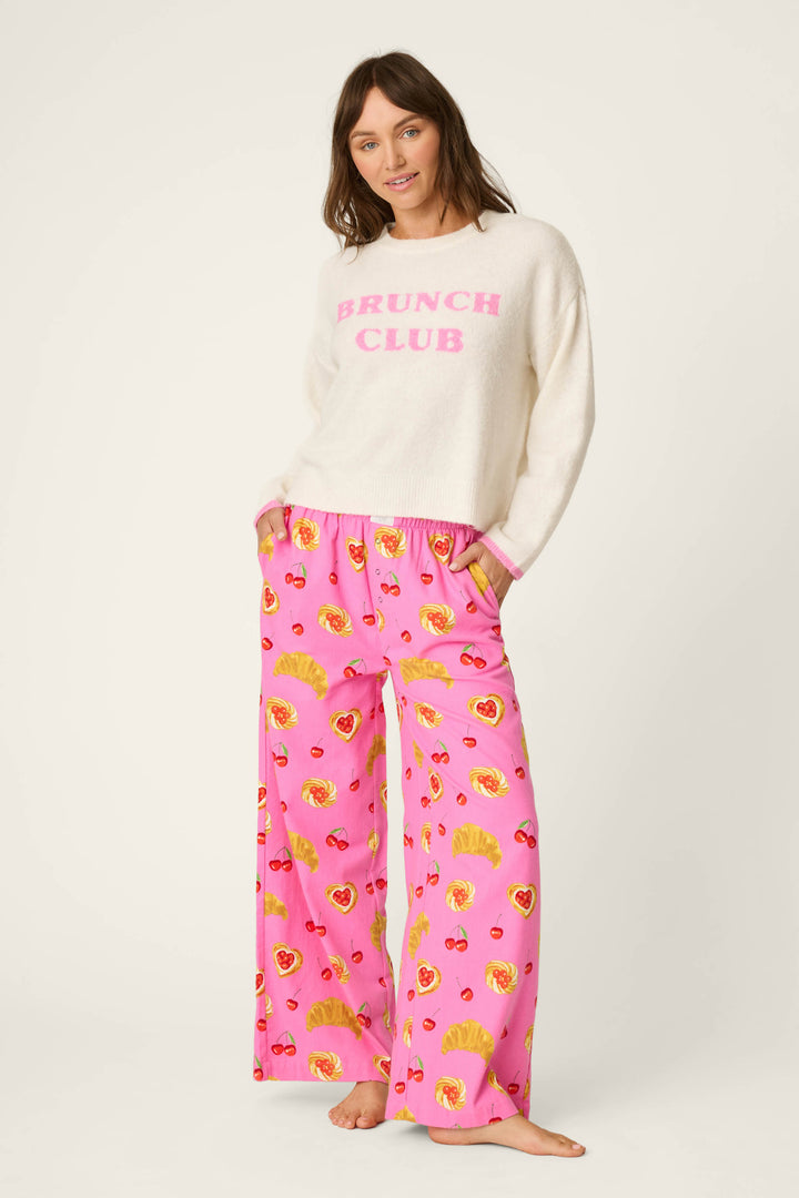 A woman stands barefoot wearing a white sweater with “BRUNCH CLUB” in pink letters and loose, bright pink Sweet Cherry Tart Print Pajama Pants by P.J. Salvage featuring a playful breakfast food print.