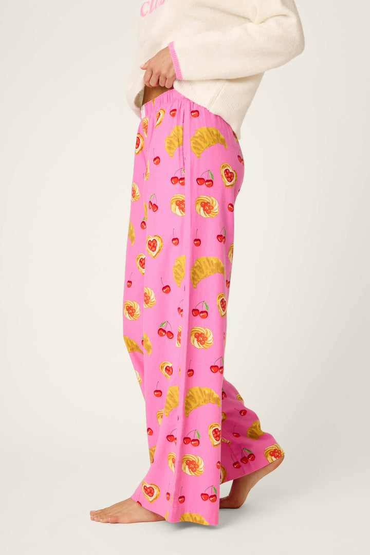 A person is wearing P.J. Salvage's Sweet Cherry Tart Print Pajama Pants, featuring playful croissant, cherry, and heart motifs, paired with a white sweater. They are barefoot and standing against a plain background.