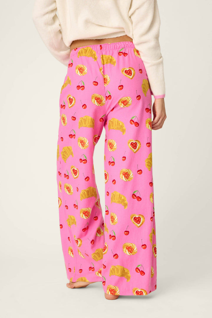 A person is standing barefoot and facing away, wearing P.J. Salvage Sweet Cherry Tart Print Pajama Pants featuring a playful pattern of croissants, cherries, and heart-shaped pastries. The upper body is partially visible in a cream sweater.