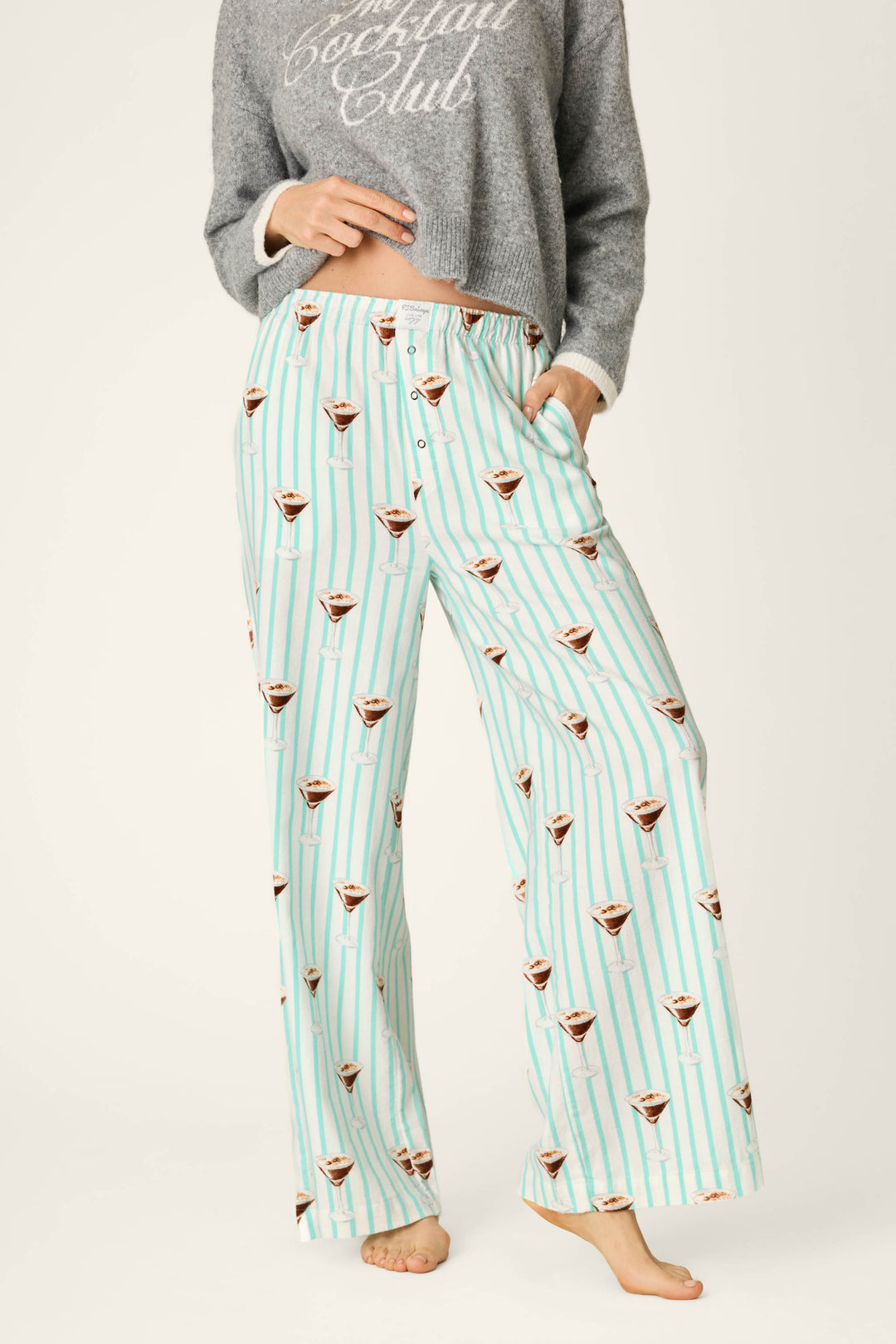 A person wearing P.J. Salvage Espresso Martini Print Flannel Pajama Pants, featuring a light blue and white striped design with espresso martini glass prints, paired with a gray cropped sweatshirt that says “Cocktail Club.” The person is barefoot and standing on a light background.