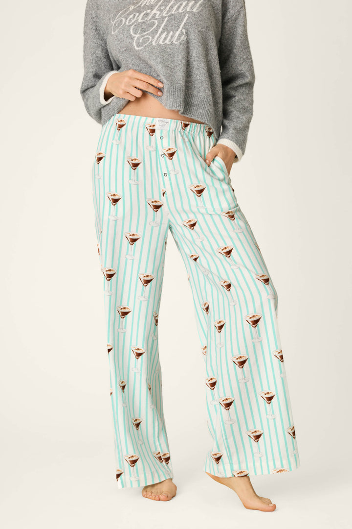 A person wearing P.J. Salvage Espresso Martini Print Flannel Pajama Pants, featuring a light blue and white striped design with espresso martini glass prints, paired with a gray cropped sweatshirt that says “Cocktail Club.” The person is barefoot and standing on a light background.