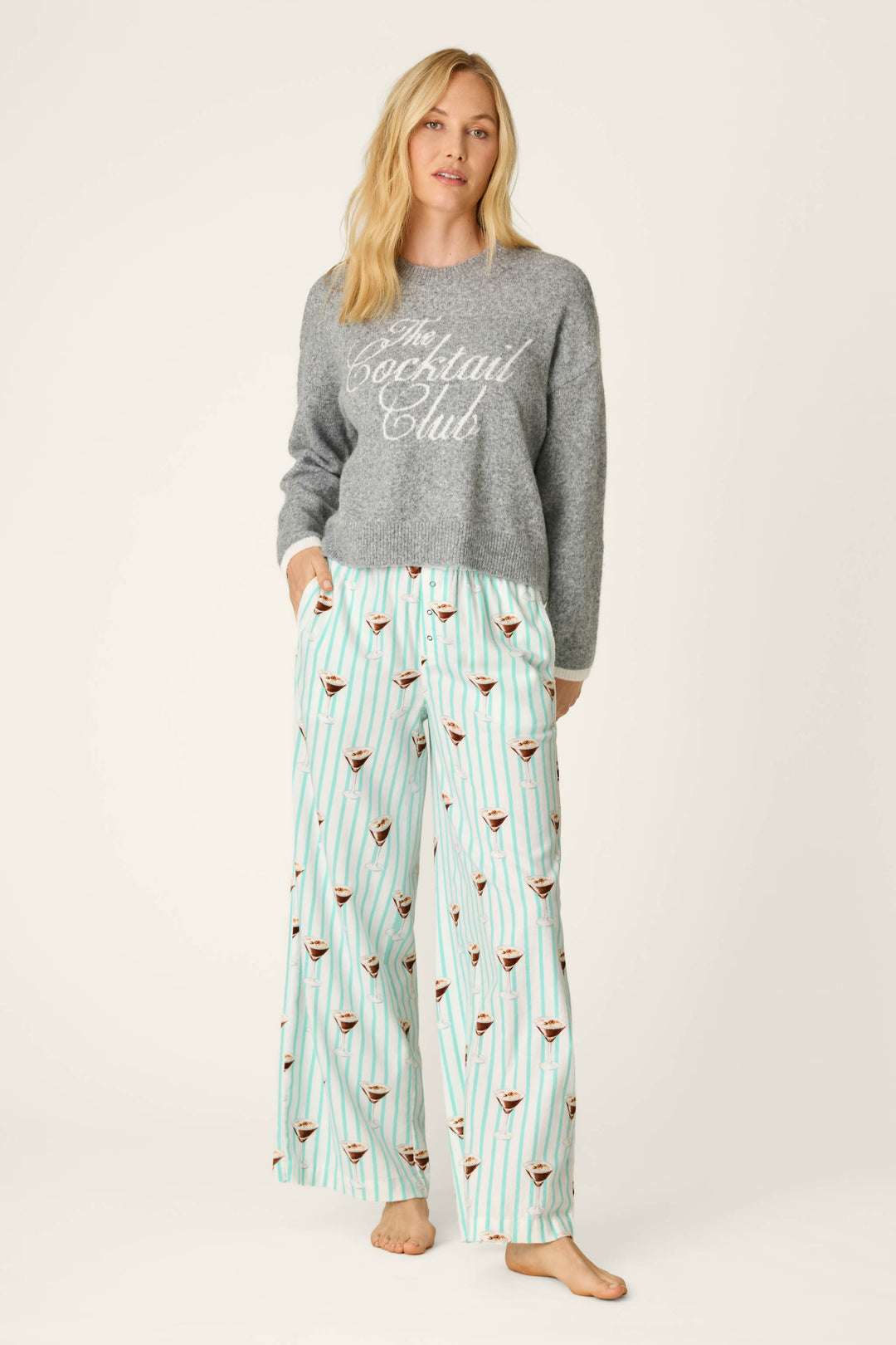 A woman wearing a grey sweater that says The Cocktail Club and P.J. Salvage Espresso Martini Print Flannel Pajama Pants, standing barefoot against a plain background.