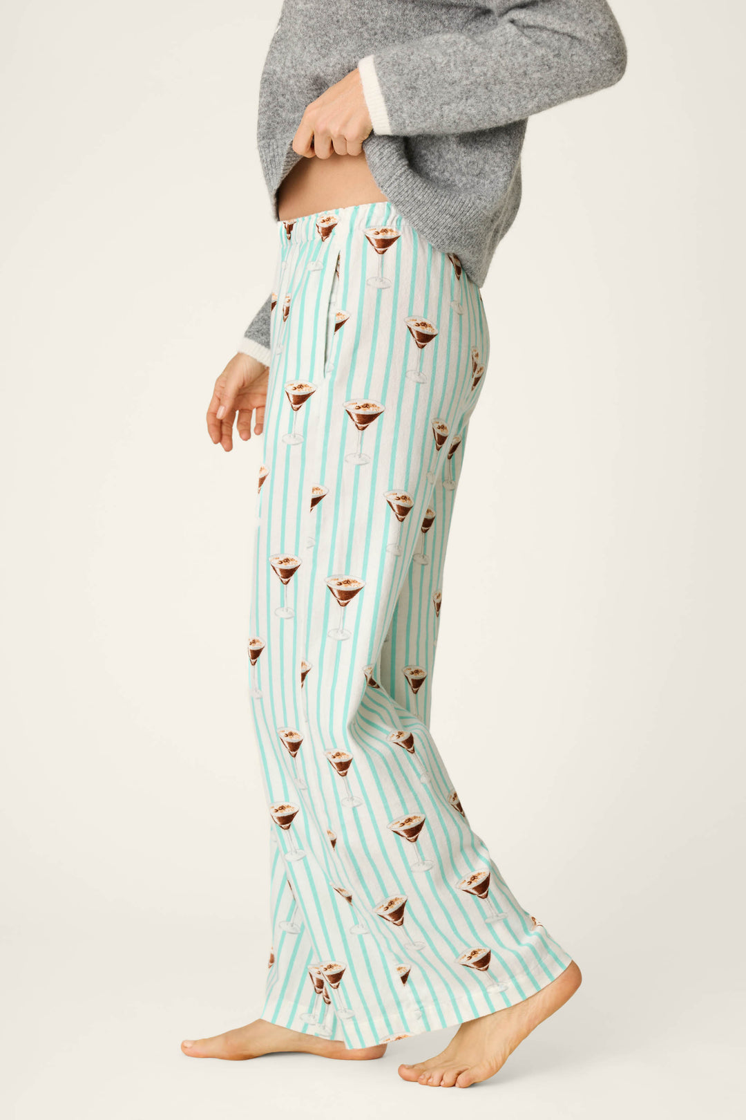 A person wearing P.J. Salvage Espresso Martini Print Flannel Pajama Pants featuring light blue stripes and a playful martini glass design, paired with a gray sweater and standing barefoot against a plain background.