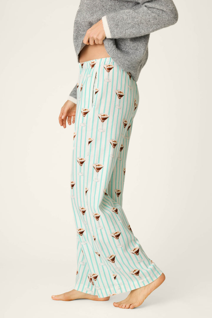 A person wearing P.J. Salvage Espresso Martini Print Flannel Pajama Pants featuring light blue stripes and a playful martini glass design, paired with a gray sweater and standing barefoot against a plain background.