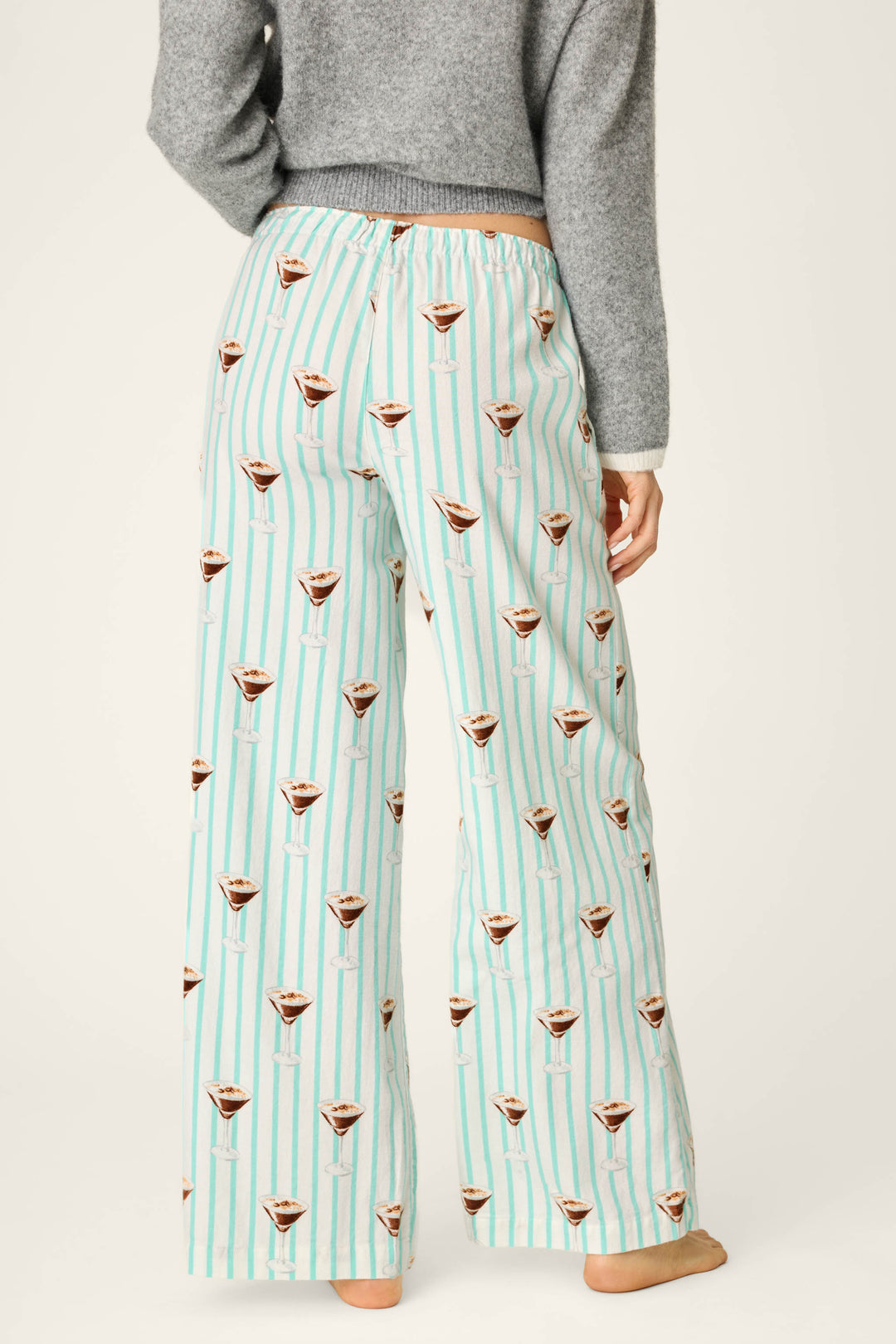 A person is shown from the back, standing barefoot and wearing P.J. Salvage Espresso Martini Print Flannel Pajama Pants in a light blue and white striped pattern with martini glass prints, paired with a gray long-sleeve top.
