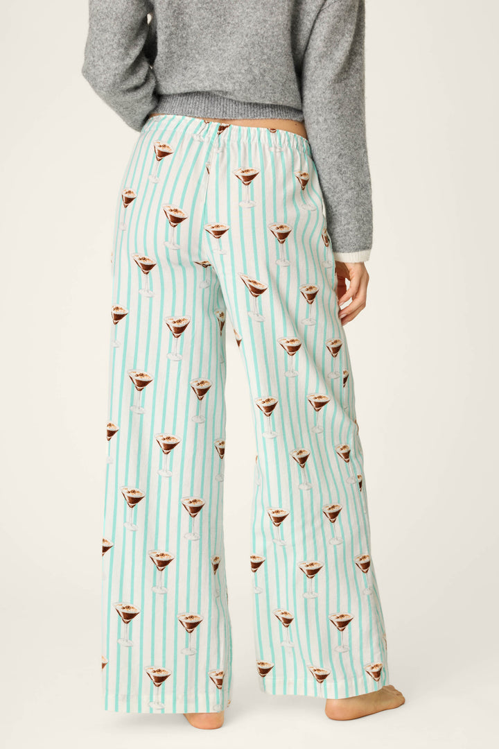 A person is shown from the back, standing barefoot and wearing P.J. Salvage Espresso Martini Print Flannel Pajama Pants in a light blue and white striped pattern with martini glass prints, paired with a gray long-sleeve top.
